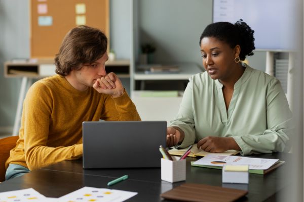 Two peopel sitting at a table working together there are laptops, papers and stationery on the table