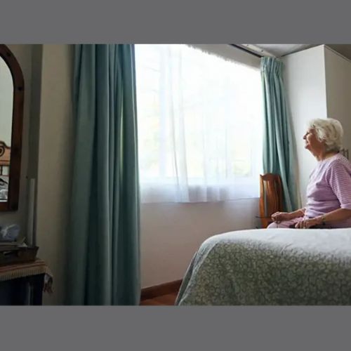 elderly lady sitting on a bed, looking out of the window