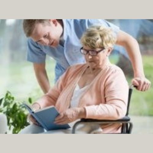 elderly lady in a wheel chair reading a book being assisted by her carer