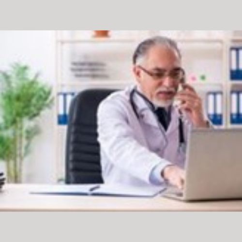 A doctor sitting at his desk speaking on the phone, looking at a laptop