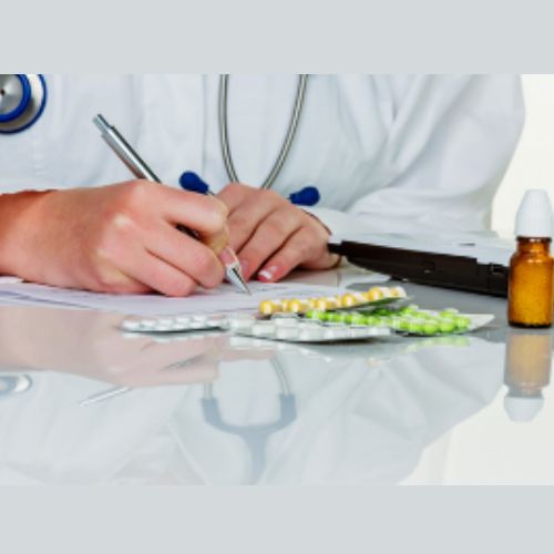 Close up of healthcare professional sitting at a desk writing, surrounded by medications  on the desk