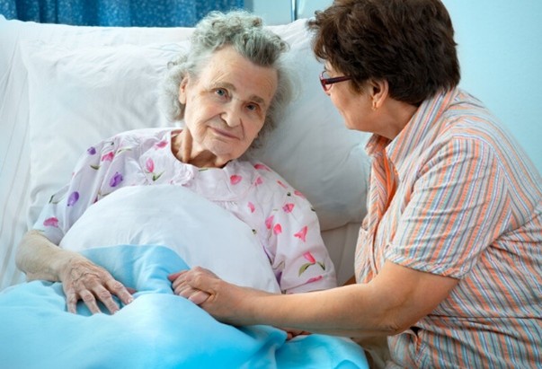elderly women laying on a hospital bed her hand hold by another woman aside